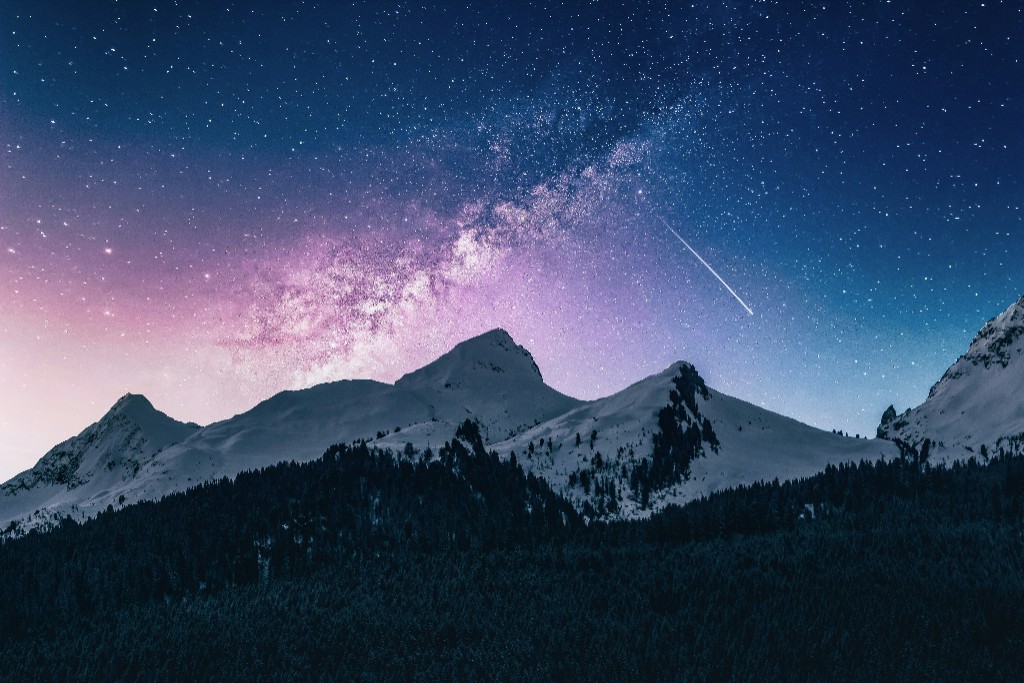 Snow-capped mountain peaks under a starry Milky Way night sky, with a dark forest in the foreground